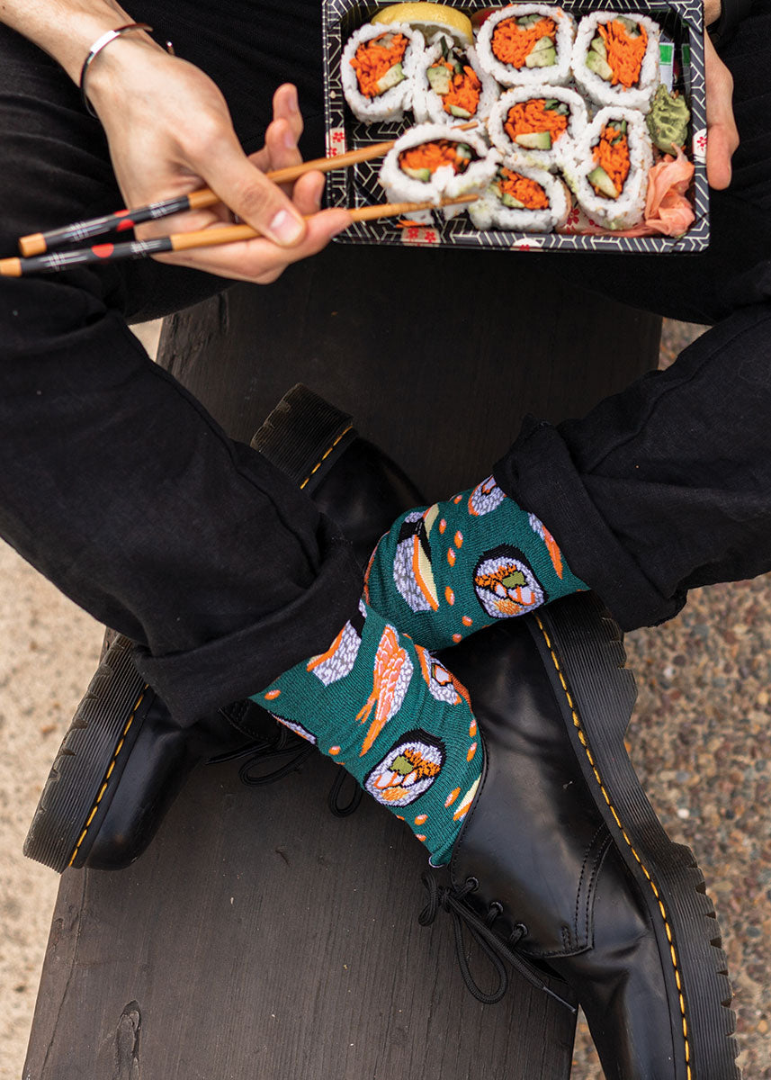 A model wearing sushi-themed novelty socks sits cross-legged with a plate of sushi in their lap. 