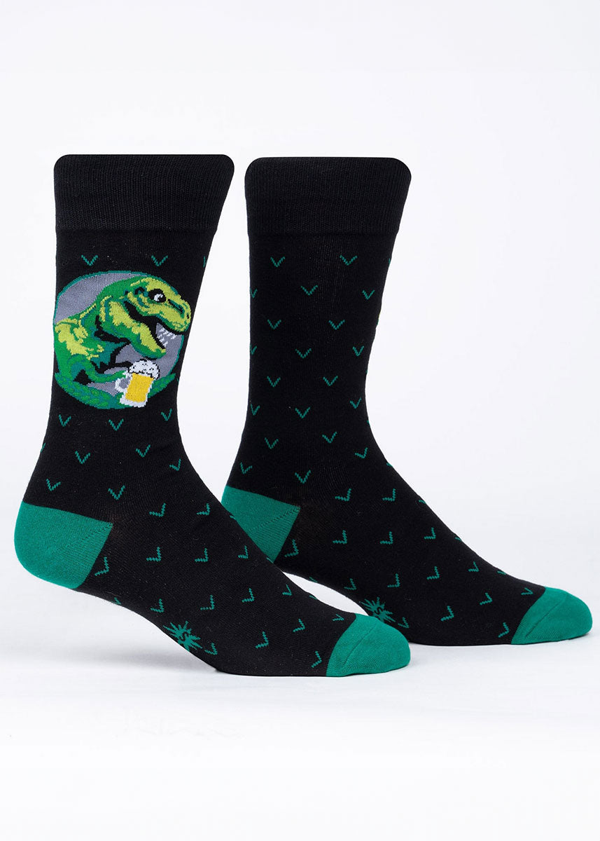 Black novelty socks for men with a dark green heel and toe featuring a green T-Rex holding a pint of beer against an allover geometric pattern.