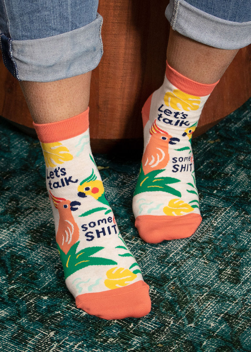 A model wearing ankle socks featuring parrots and tropical plants that say &quot;Let&#39;s Talk Some Shit&quot; poses against a teal carpet.