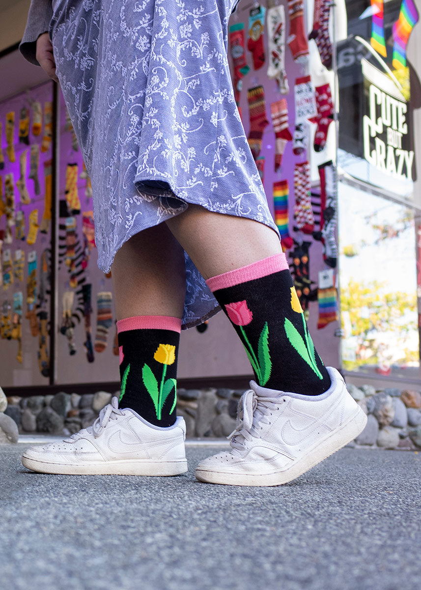 A model wearing tulip-themed novelty socks, a floral skirt and white sneakers poses in front of a sock store window.