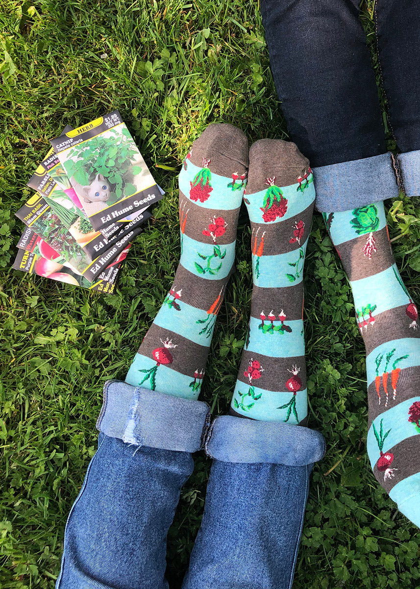 Two models wearing vegetable garden-themed novelty socks pose seated together in the grass next to seed packets.