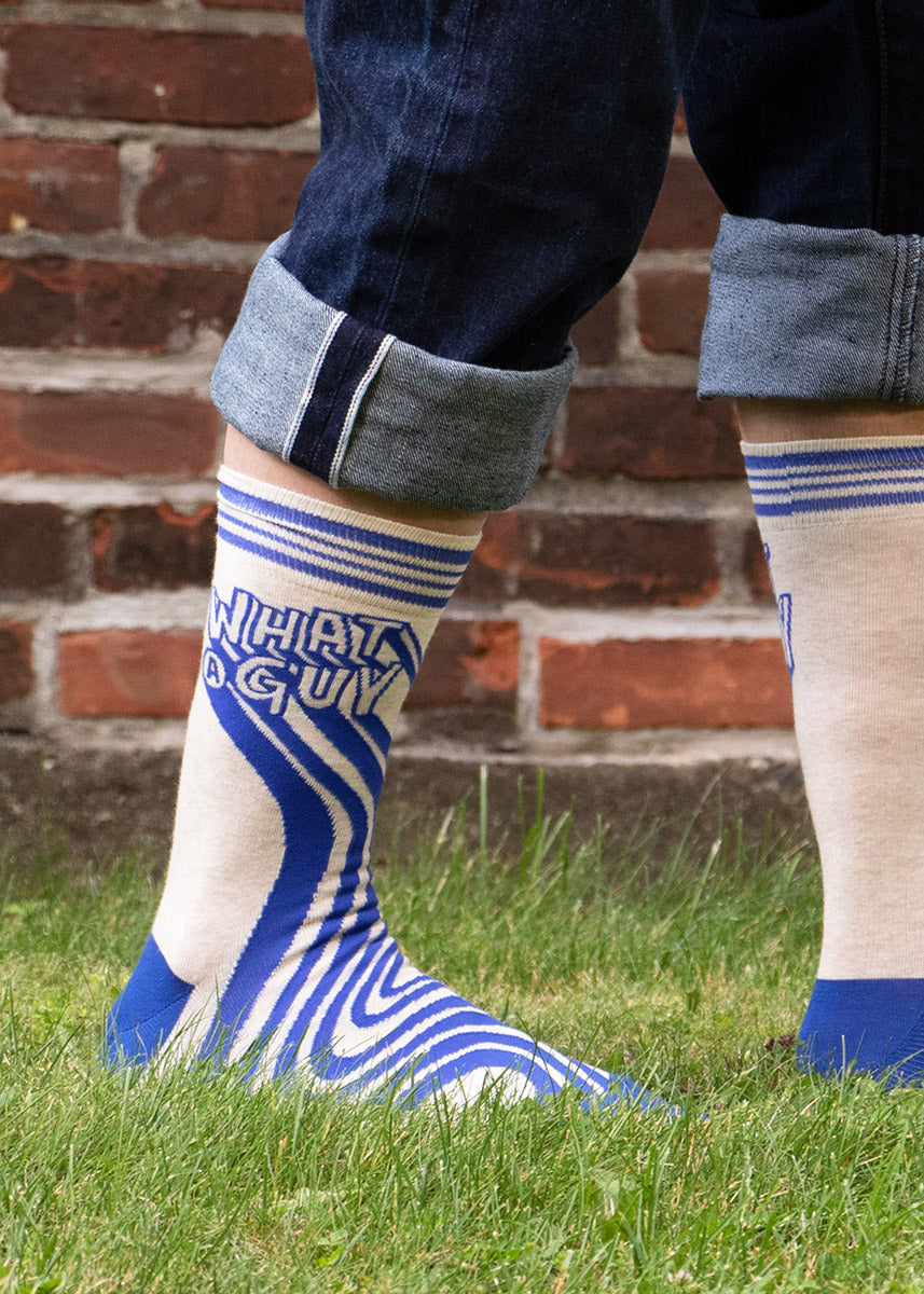 A model wearing novelty socks that read &quot;What a Guy&quot; poses in the grass against a red brick wall.