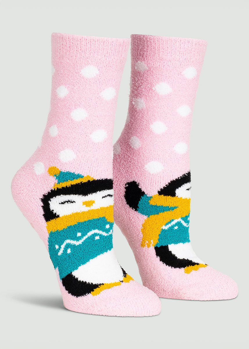Light pink fuzzy slipper socks for women with white polka dots featuring a penguin dressed up in a teal sweater, yellow scarf, and hat.