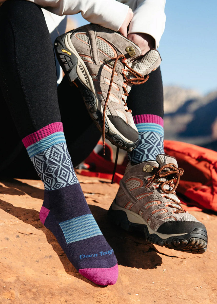 A model wearing blue and purple geometric hiking socks poses outside putting on hiking boots.