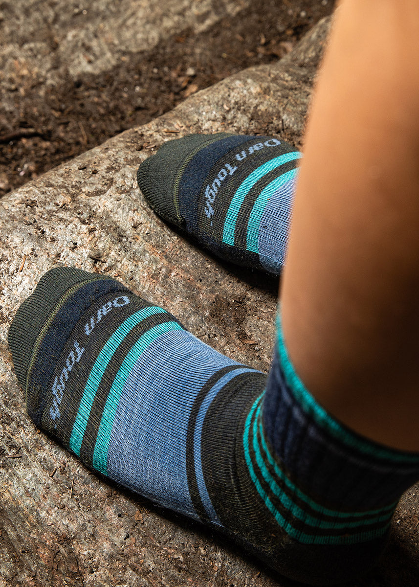 A model poses outside on a rock wearing blue and dark olive green striped wool boot socks for women.