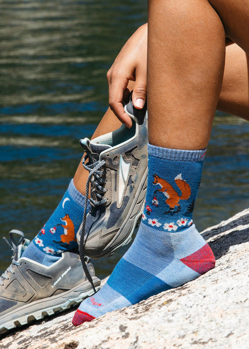 Blue hiking crew socks with red accents depict a whimsical scene of a fox carrying a lantern through a forest full of trees, flowers and small red mushrooms with a crescent moon up above.