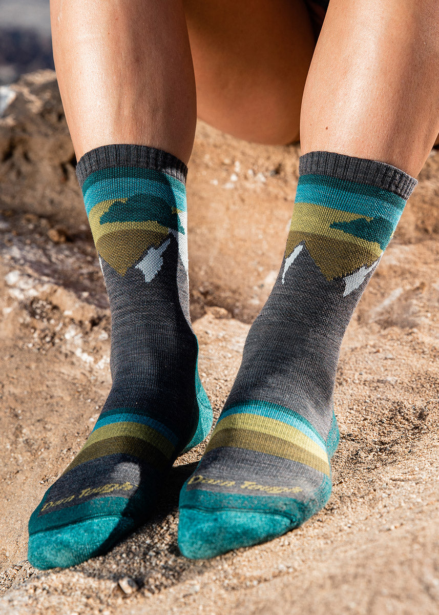 Gray hiking socks for women with snow-capped mountain peaks and a background of teal, blue, and yellow stripes.