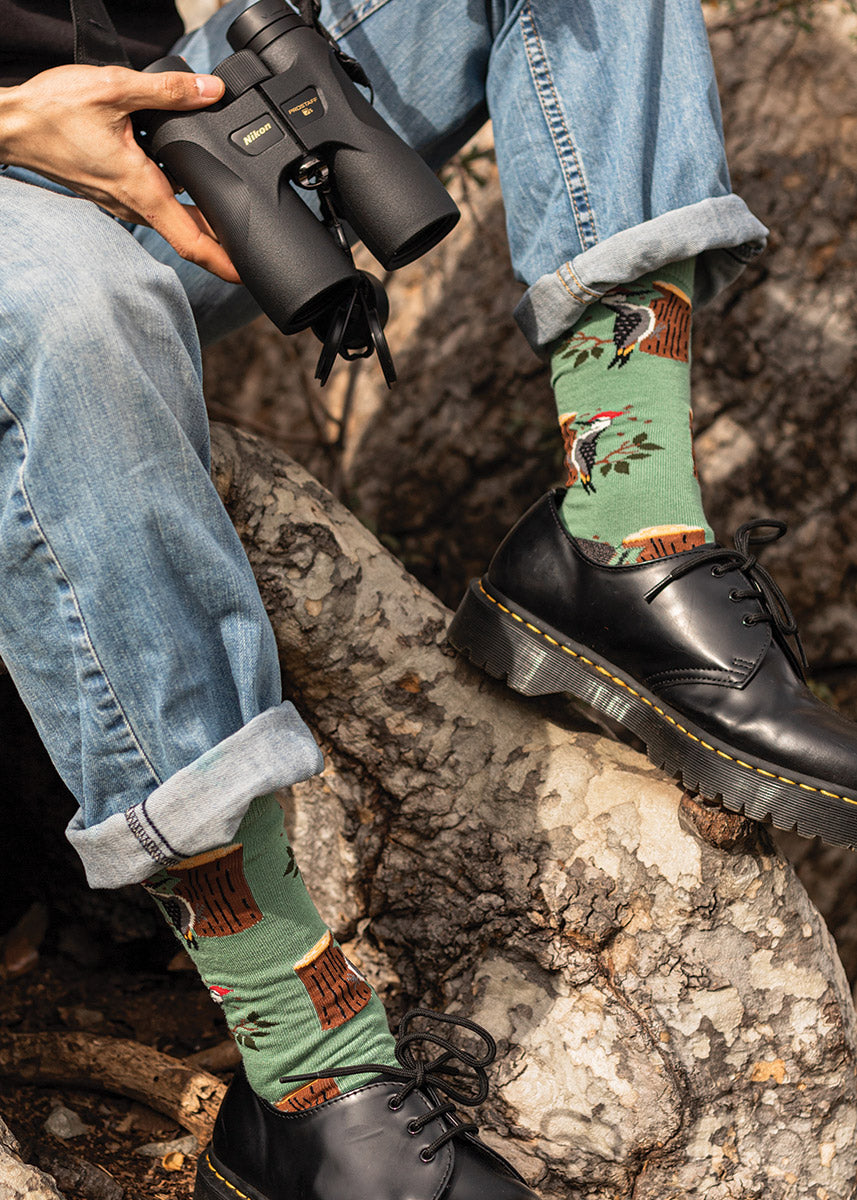 A model wearing woodpecker-themed novelty socks and holding binoculars poses outside sitting against a tree. 