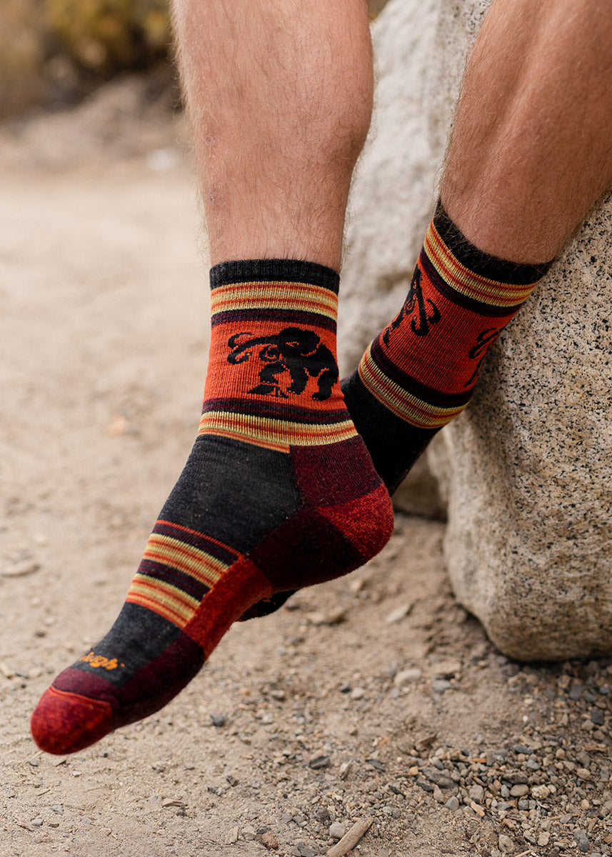 A male model wearing red and orange woolly mammoth-themed wool hiking socks poses outside sitting on a large rock.
