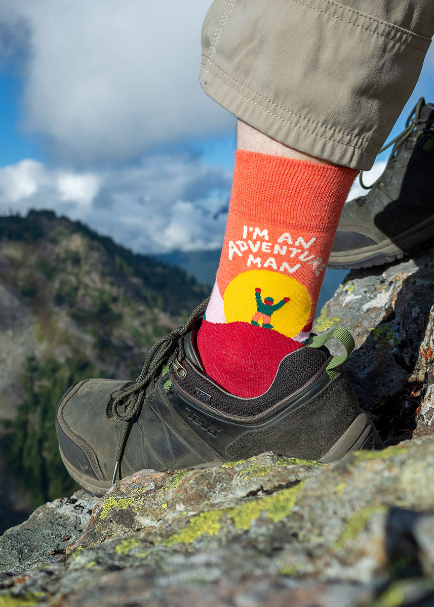 A male model wears a pair of novelty socks reading &quot;I&#39;m an adventure man&quot; with brown hiking boots while posed atop a cliffside