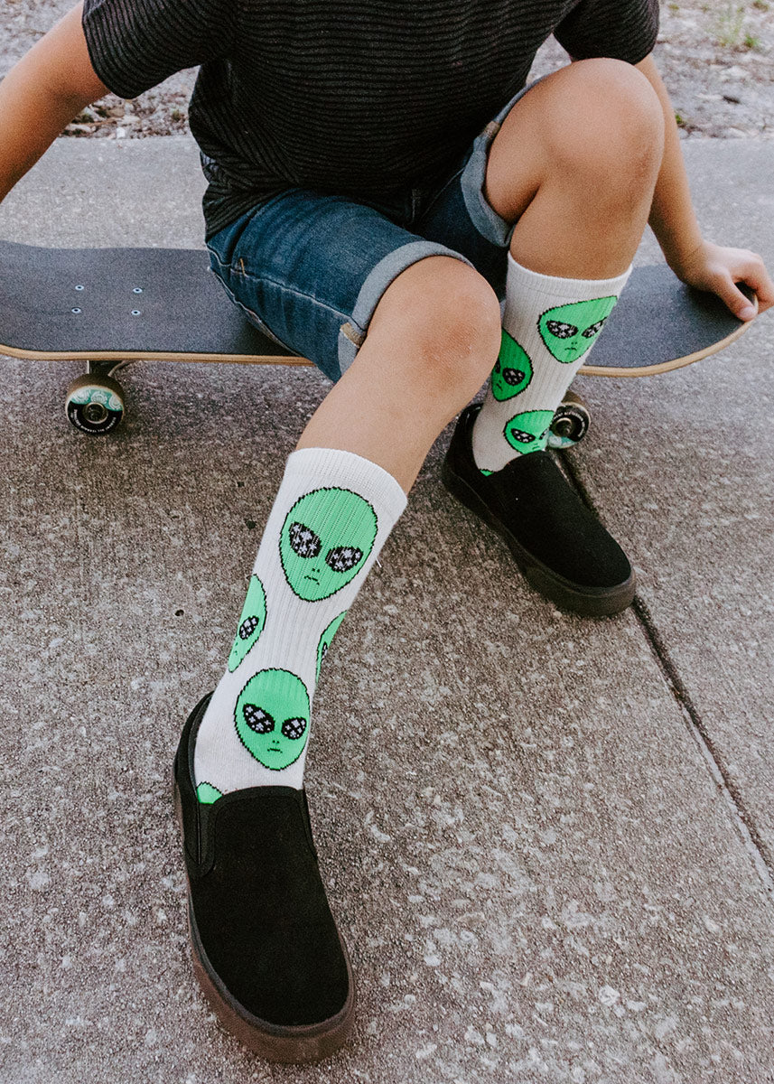 A male child model wearing alien face novelty socks and black sneakers poses seated on a skateboard.