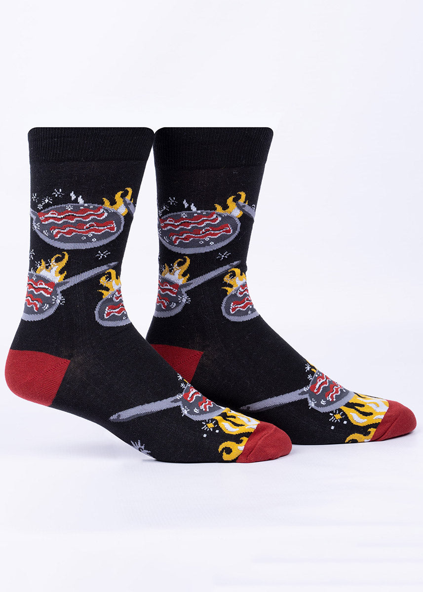 Black crew socks for men with a pattern of strips of bacon cooking in a frying pan over a high flame, accented with dark red at the heel and toe.
