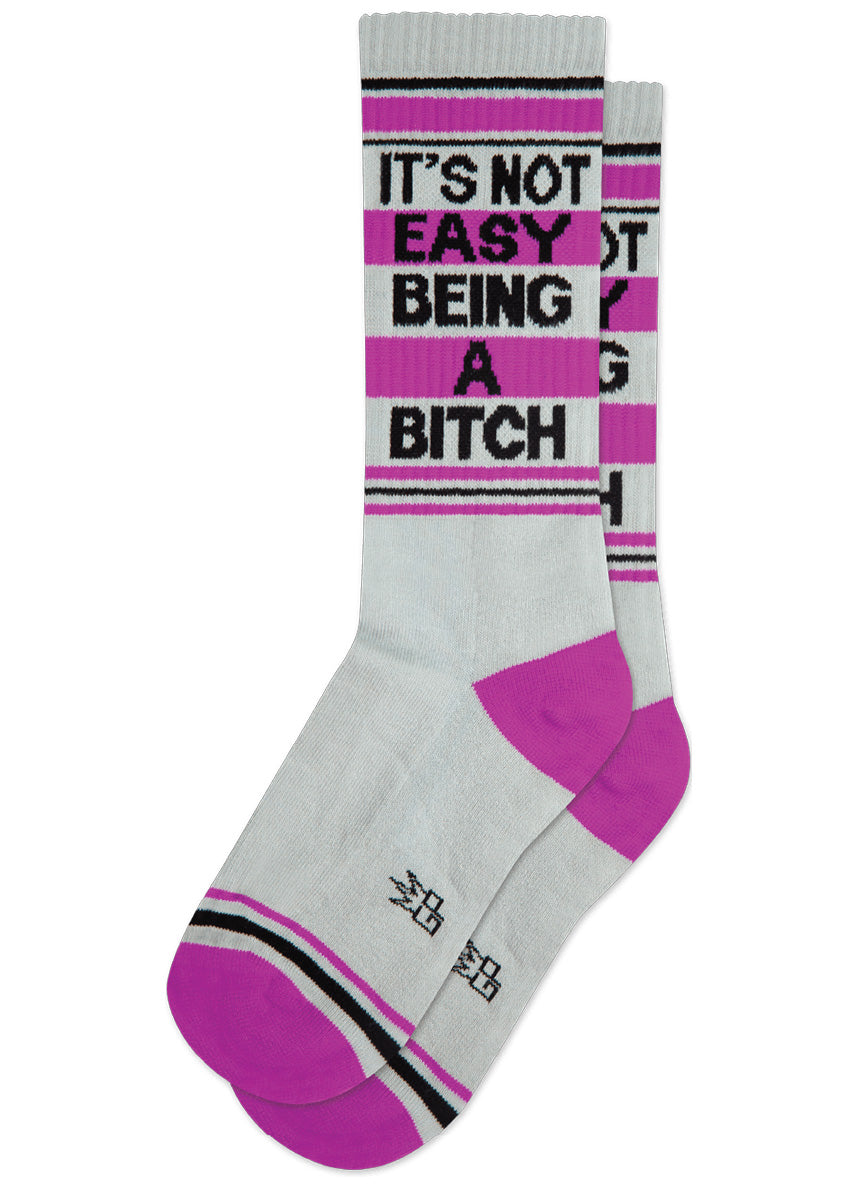 Funny retro gym socks say "It's not easy being a bitch" on a light gray background with magenta and black accents.