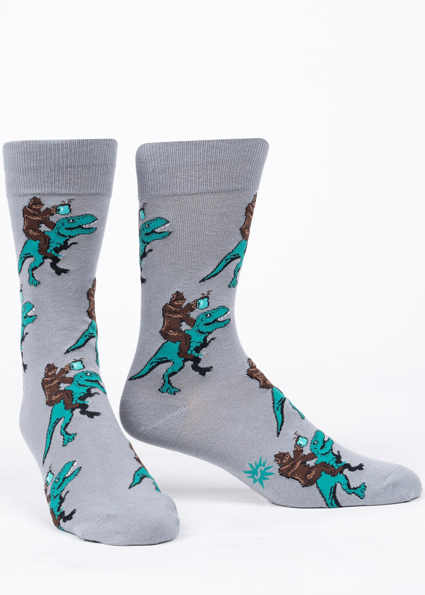 Funny socks for men show Bigfoot riding a turquoise T-Rex and holding a mug of hot coffee.