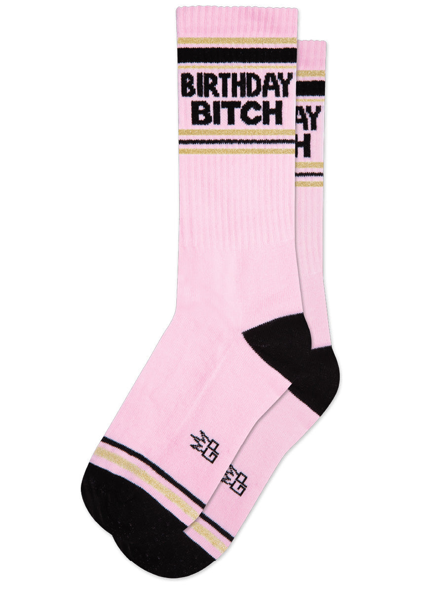 Retro athletic-style crew socks have the words “BIRTHDAY BITCH” on a pink background with stripes in black and gold sparkle.