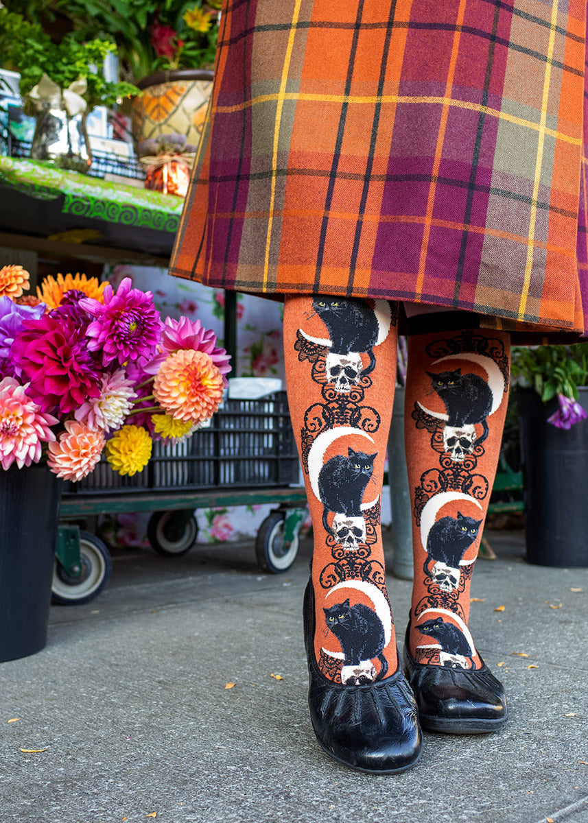 A female model wearing orange black cat-themed novelty knee socks and a plaid skirt poses in front of flowers.