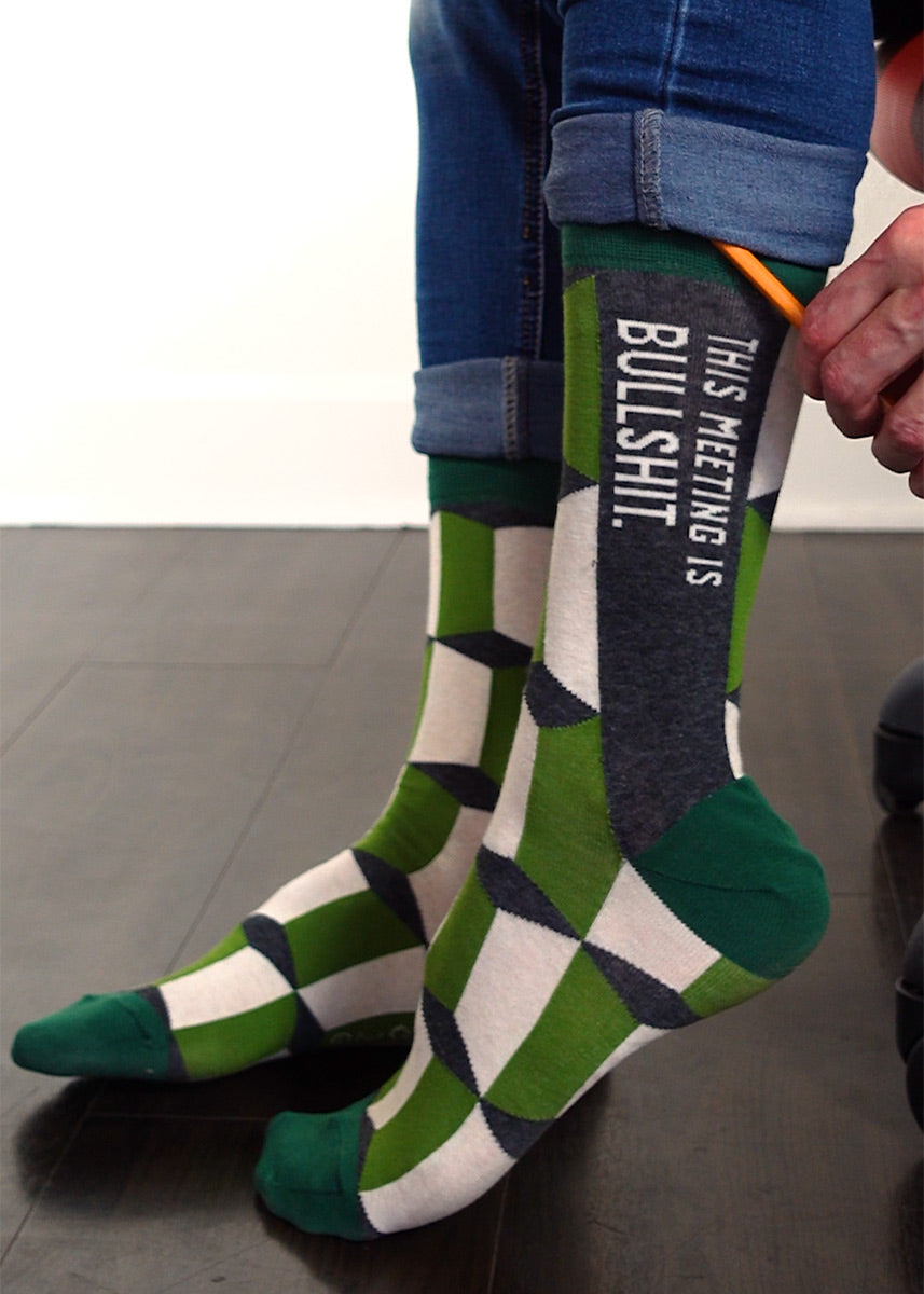 Funny swear word men's socks that say "This Meeting is Bullshit" on a green patterned background.