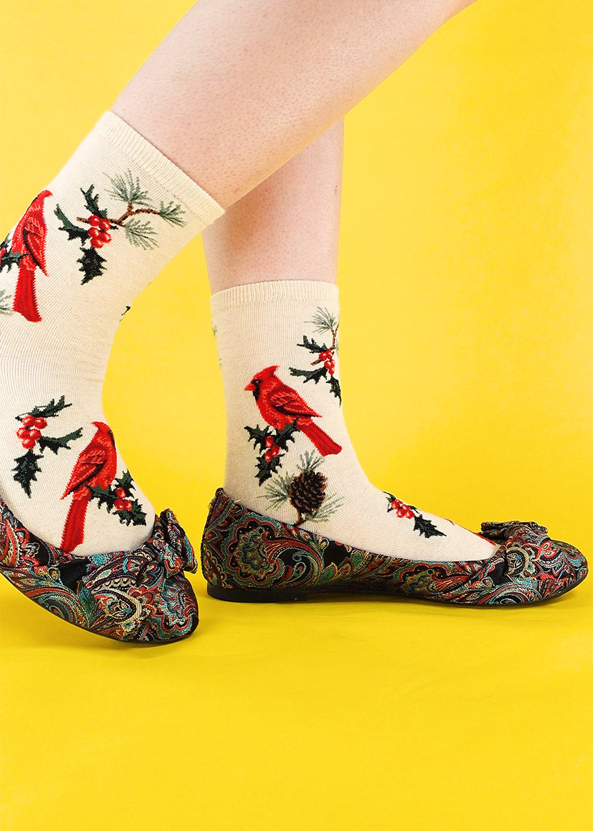 A model stands with a bright yellow background, in a pair of creme colored socks decorated with red cardinals, holly leaves, and pine cones in a pair of patterned ballet flats. 