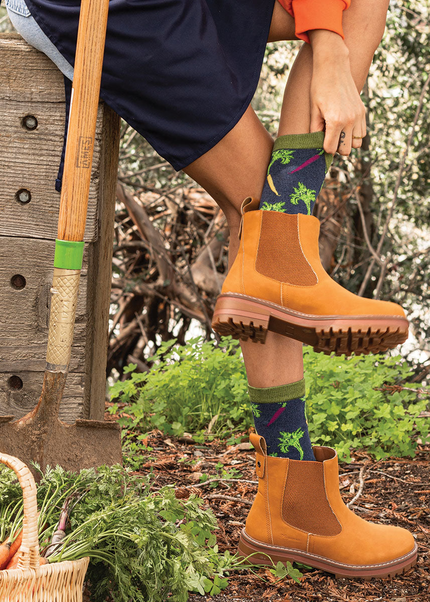 A model wearing carrot-themed novelty socks poses outside in a garden with a shovel and a basket of carrots.