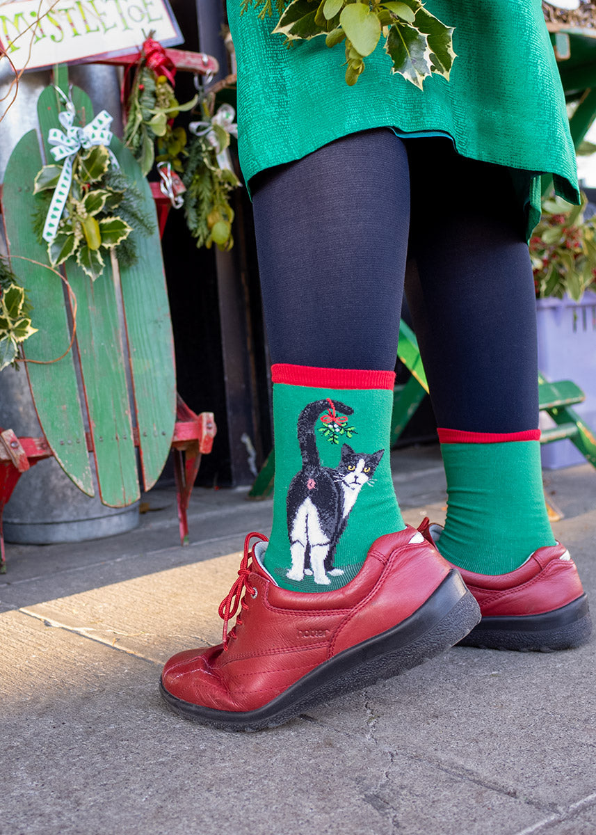 A female model wearing Christmas cat-themed novelty socks and red shoes poses next to mistletoe.