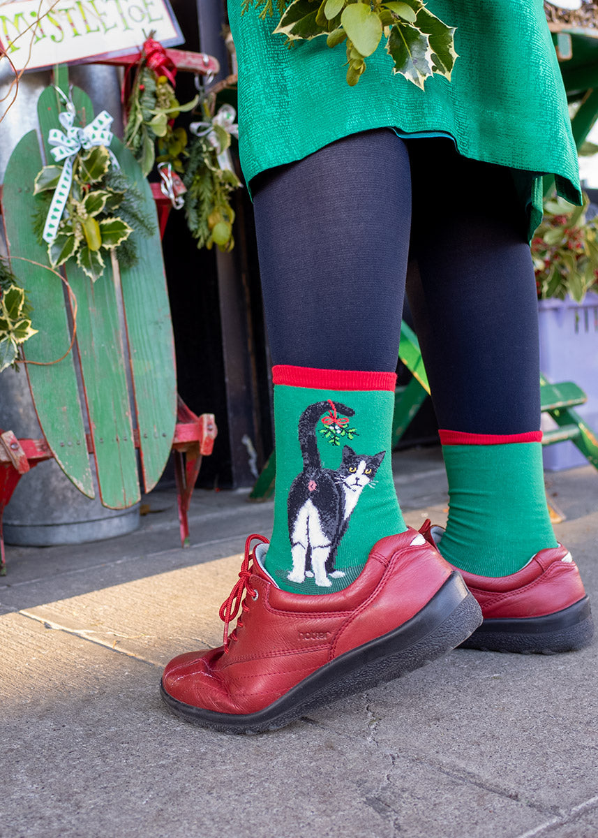 Green women's crew-length Christmas socks with red accents depict Tuxedo cats showing off their behind with a sprig of mistletoe hanging from their tail, giving a cheeky look from over their shoulder.