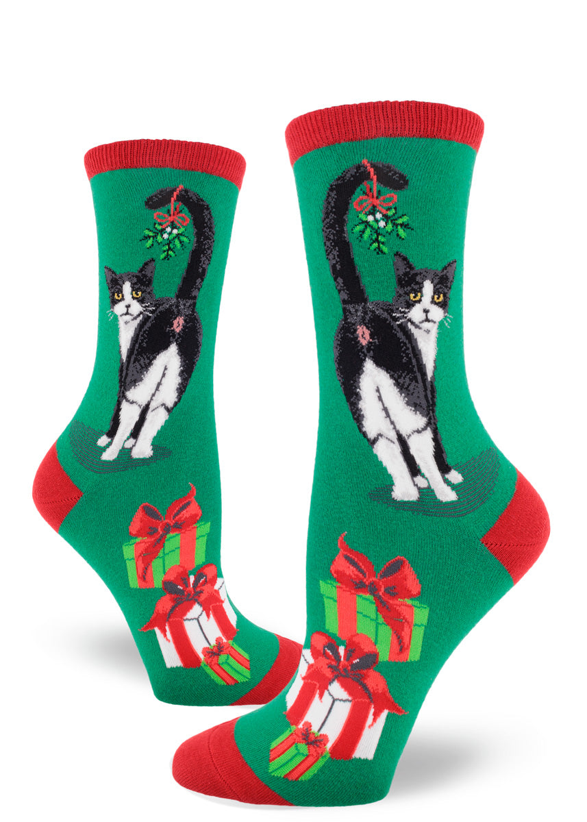 Green women's crew-length Christmas socks with red accents depict Tuxedo cats showing off their behind with a sprig of mistletoe hanging from their tail, giving a cheeky look from over their shoulder.