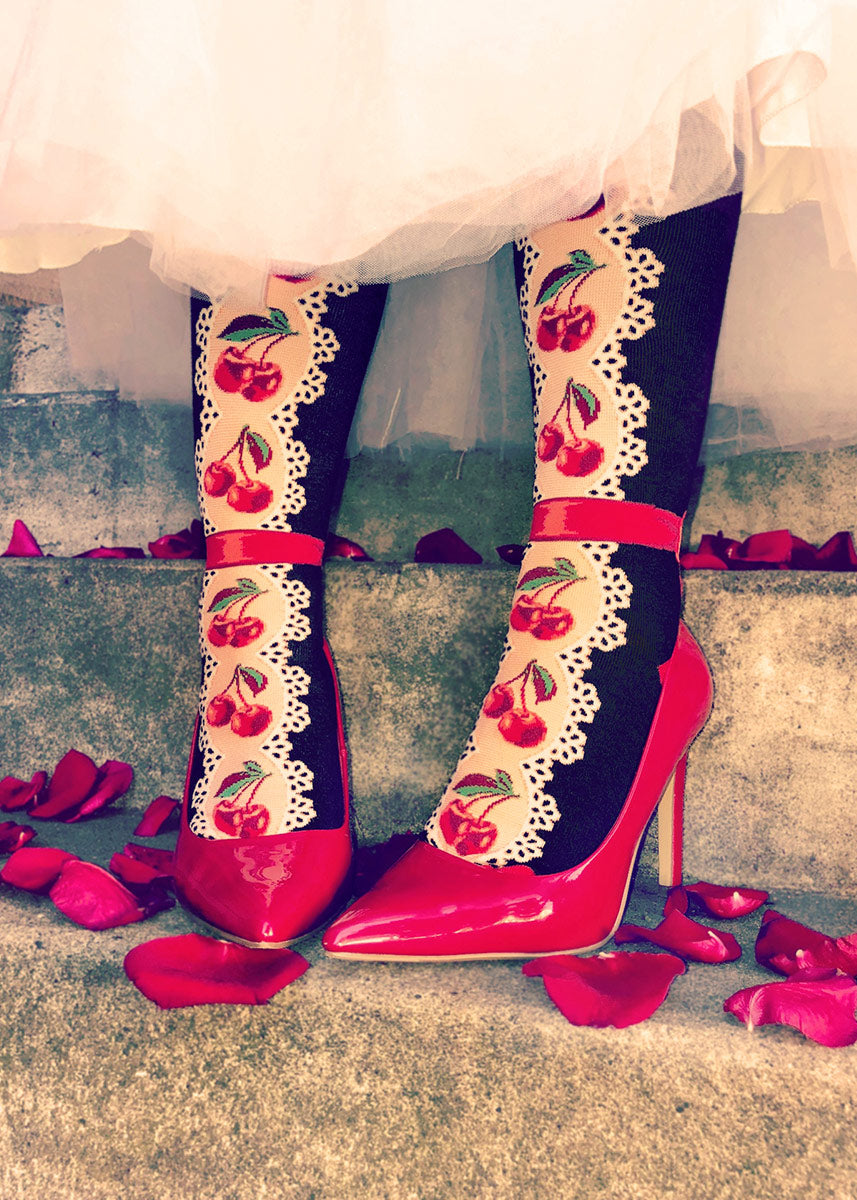 A female model wearing cherry and lace-themed knee socks and red heels poses standing on stairs covered with rose petals