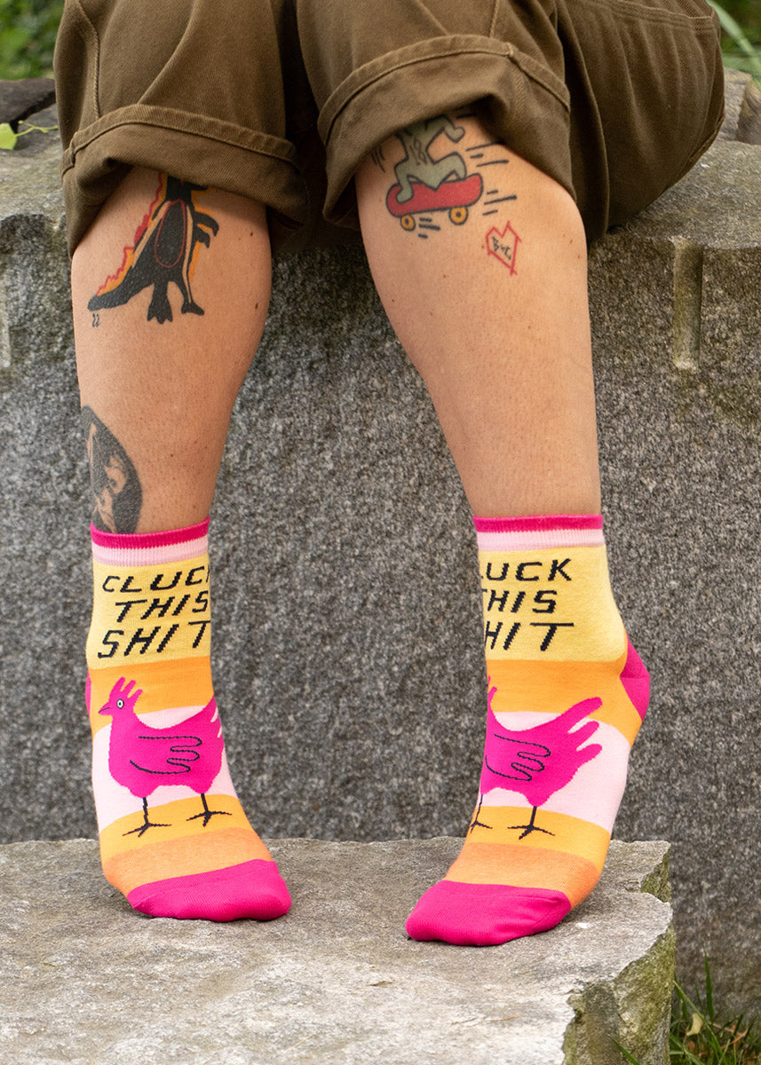 A tattooed model wearing chicken-themed novelty ankle socks and no shoes sits on a rock.
