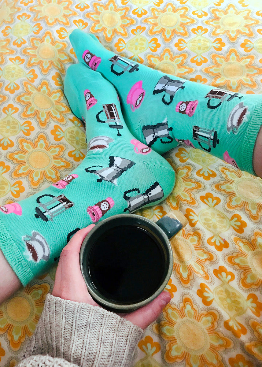 Coffee socks with a pattern of French press coffee pots, cups of coffee and to-go cups on a seafoam green background.