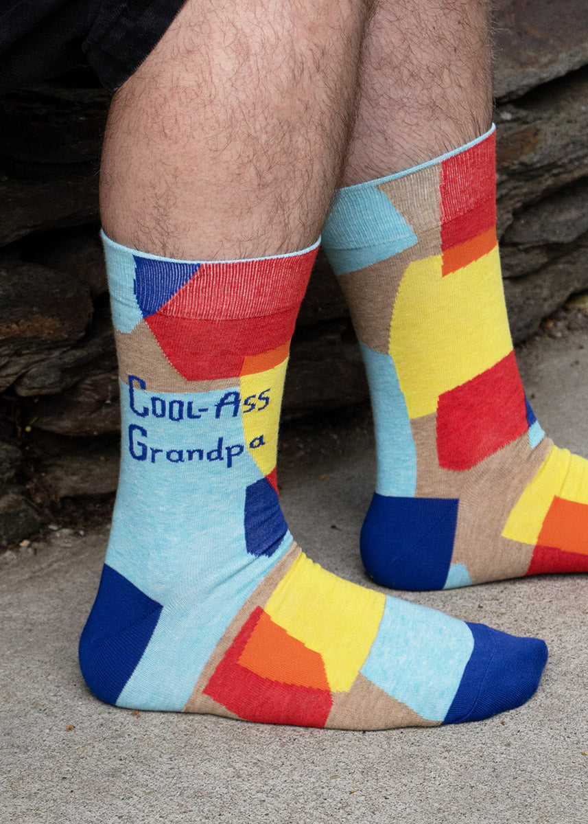 Funny crew socks for men feature yellow, red, and blue shapes on a taupe background with the words, "Cool-Ass Grandpa."