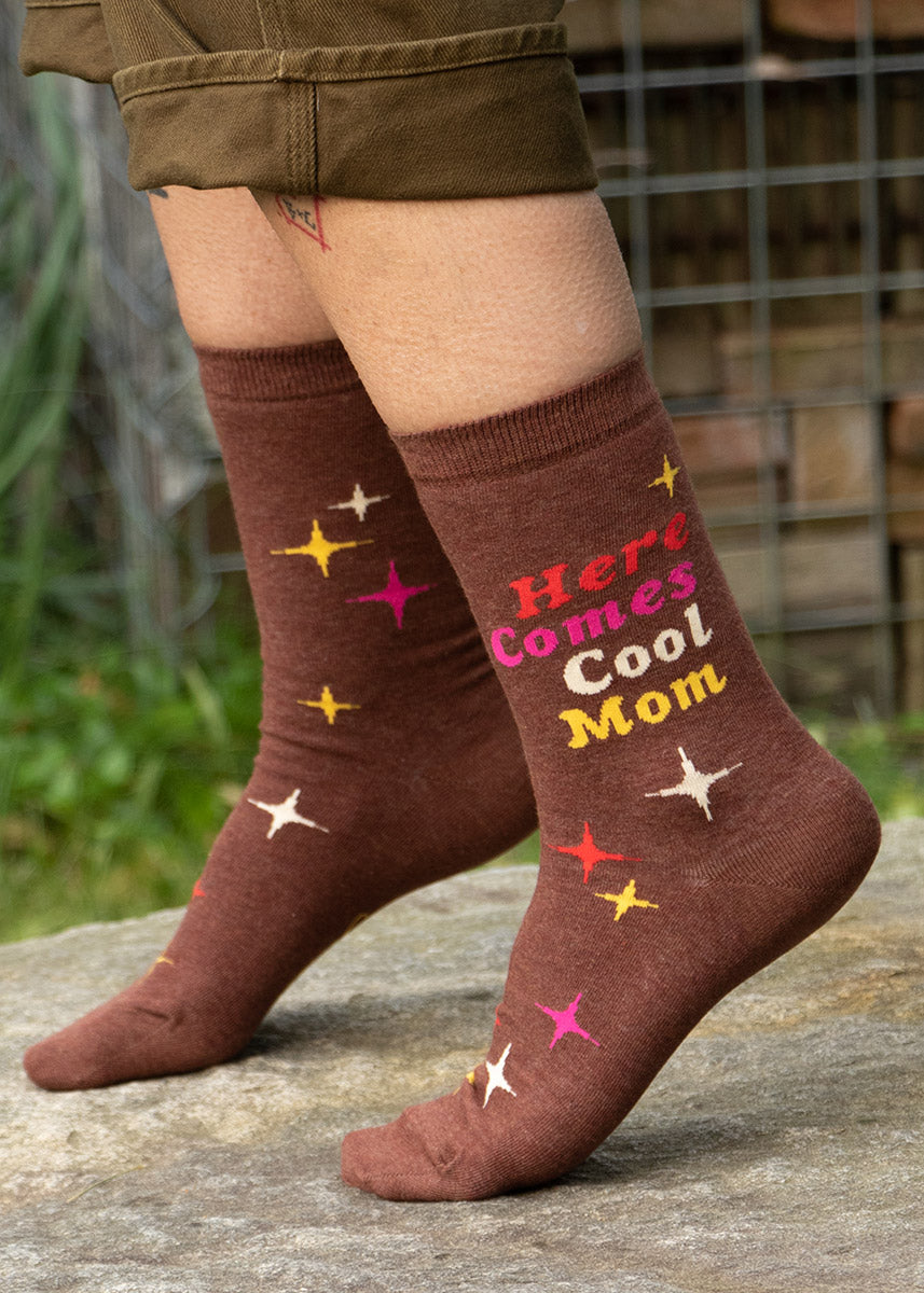 A model wearing novelty socks that read &quot;Here Comes Cool Mom&quot; poses outside on the sidewalk.