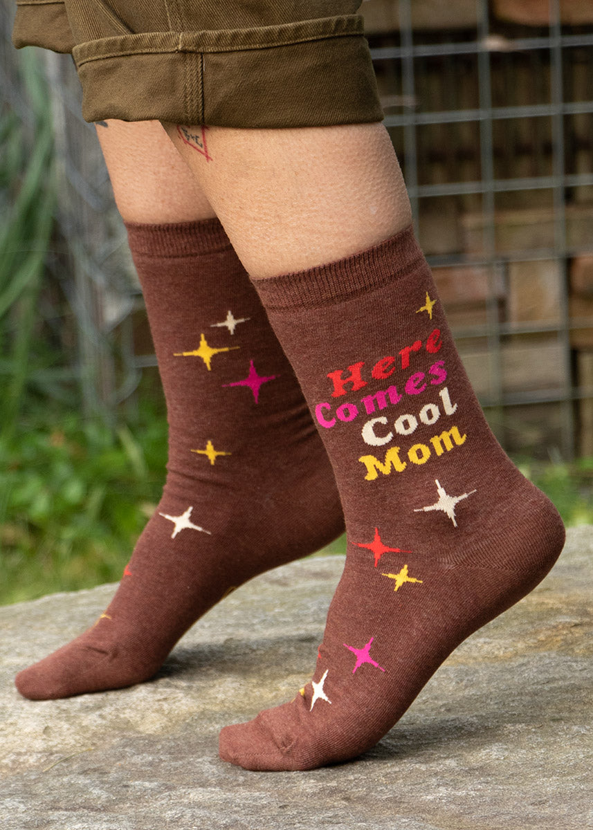 Brown crew socks that say “Here Comes Cool Mom” in colorful retro lettering surrounded by an embellishment of four-pointed atomic stars.