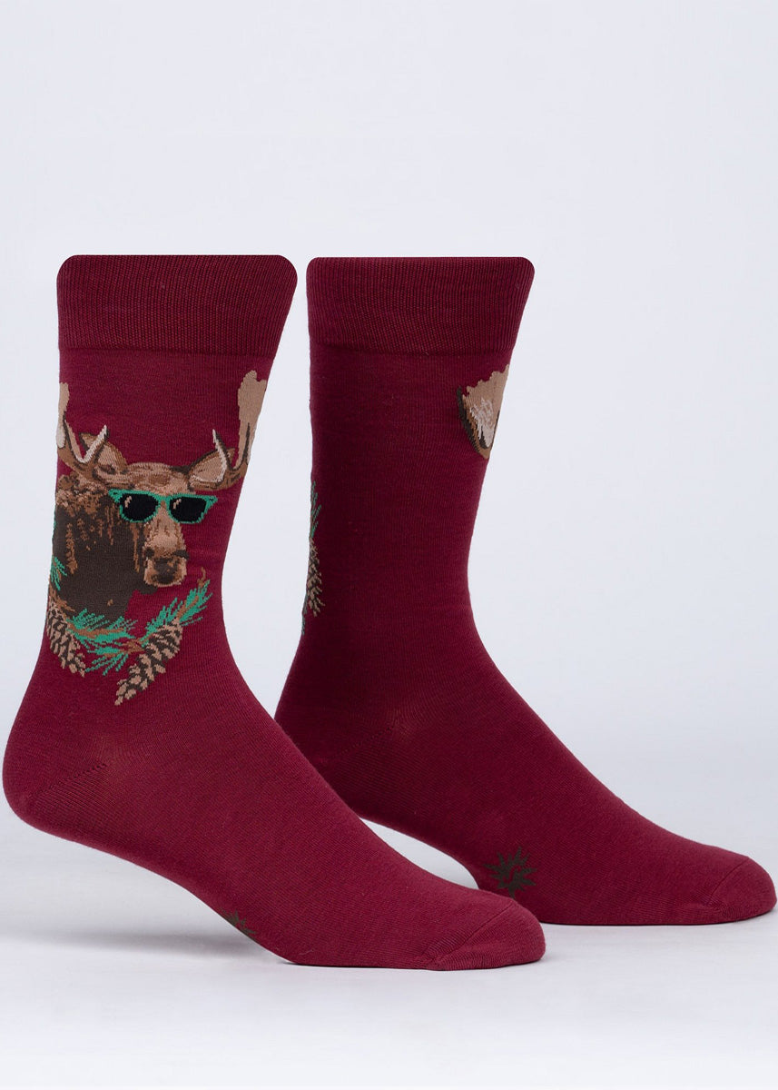 Funny animal socks for men feature a big bull moose wearing green sunglasses behind a pinecone-covered branch on a dark red background.
