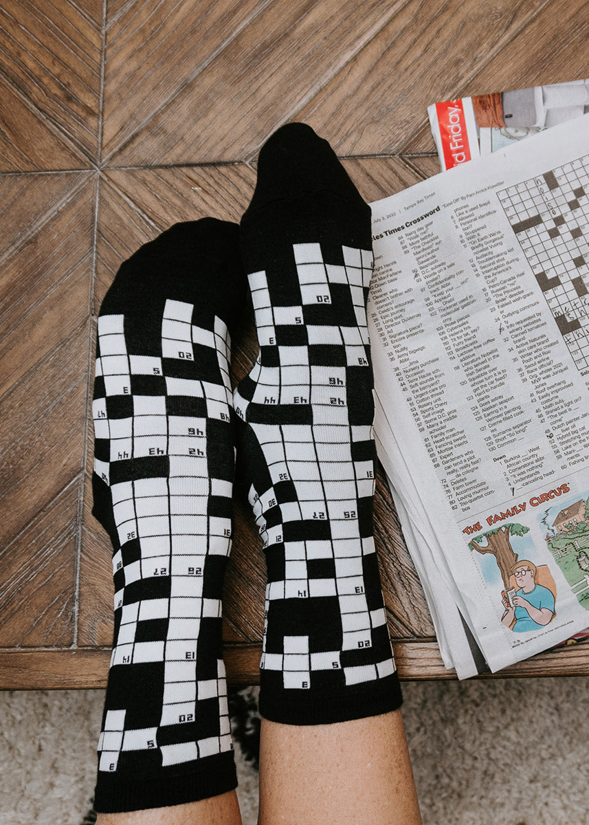 A model wearing crossword puzzle themed novelty socks poses next to a crossword page in a newspaper.