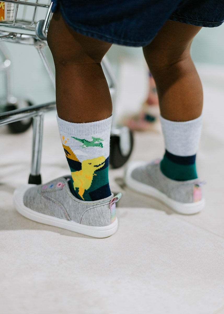 A smiling yellow T. rex walks across these dinosaur crew socks for kids, which also feature a green pterodactyl, a volcano and a palm tree.