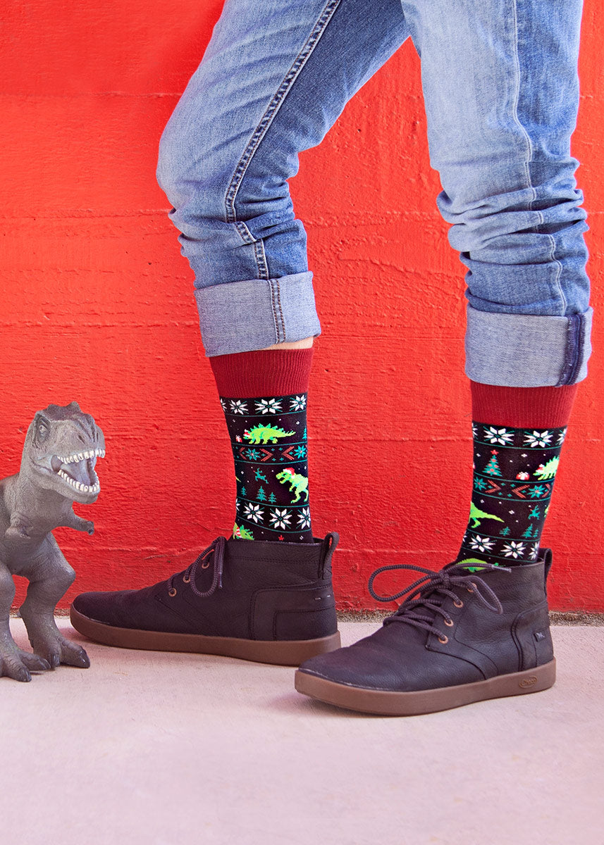 A model wearing dinosaur and Christmas-themed novelty socks poses next to a toy dinosaur against a red background.