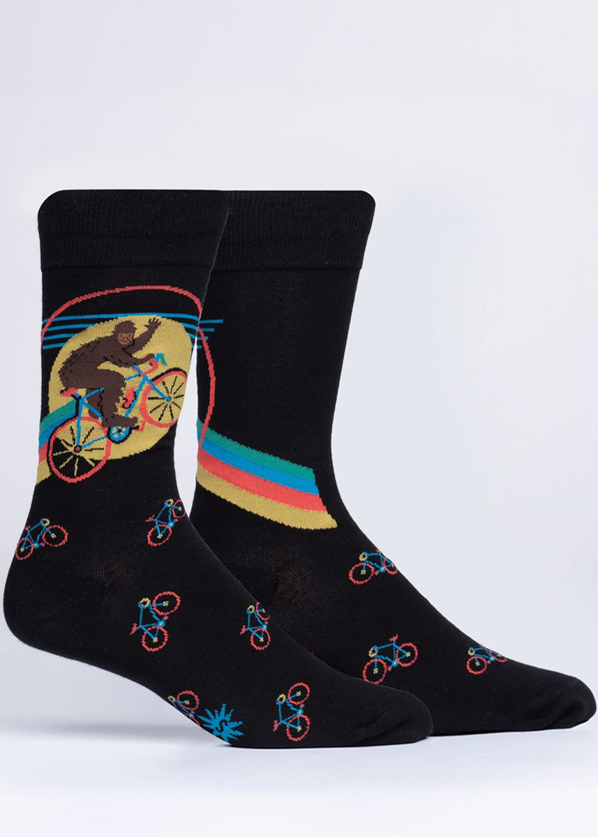 Funny crew socks for men show Sasquatch waving as he rides a bike through the sky and past the moon followed by a rainbow!