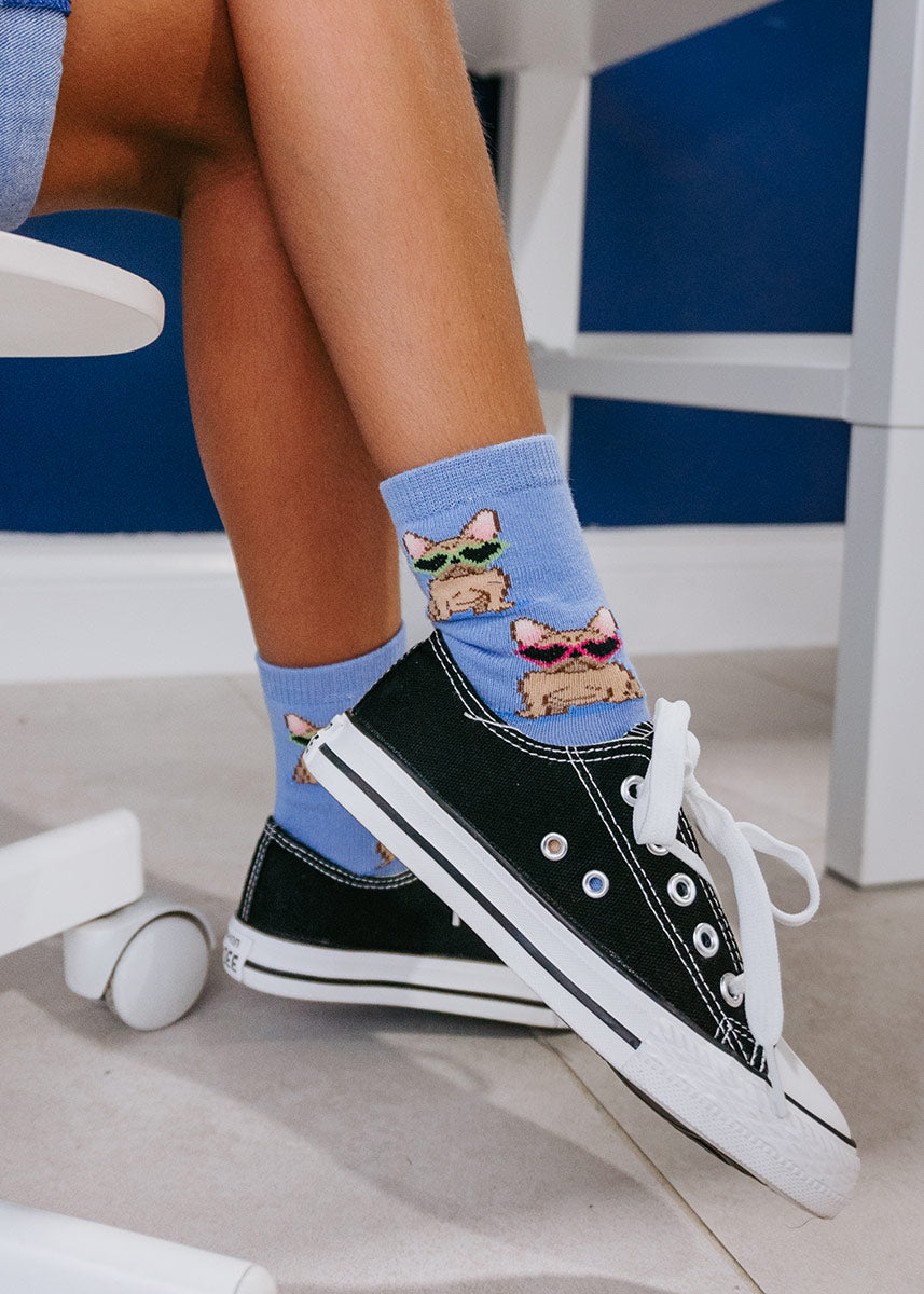 A child model wearing french bulldog-themed novelty socks and black sneakers poses seated at a desk.