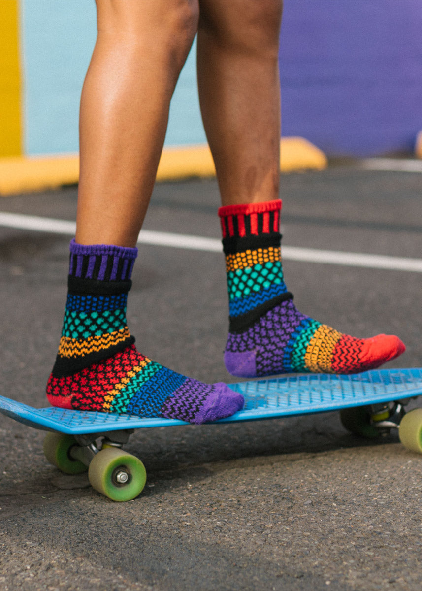 A model wearing rainbow mismatched socks poses riding on a skateboard.