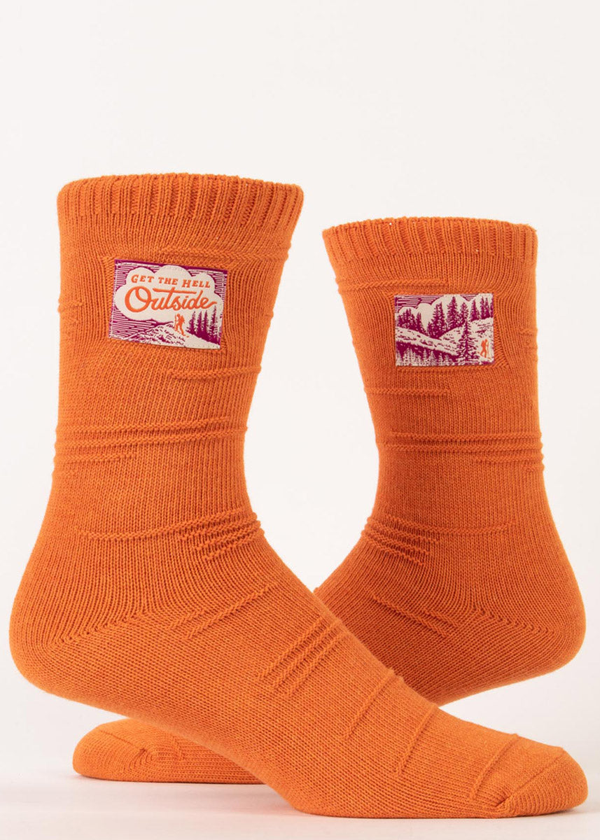 Funny nature socks feature tags showing a backpacker hiking a mountain with the words &quot;Get the hell outside&quot; on an orange knit background.