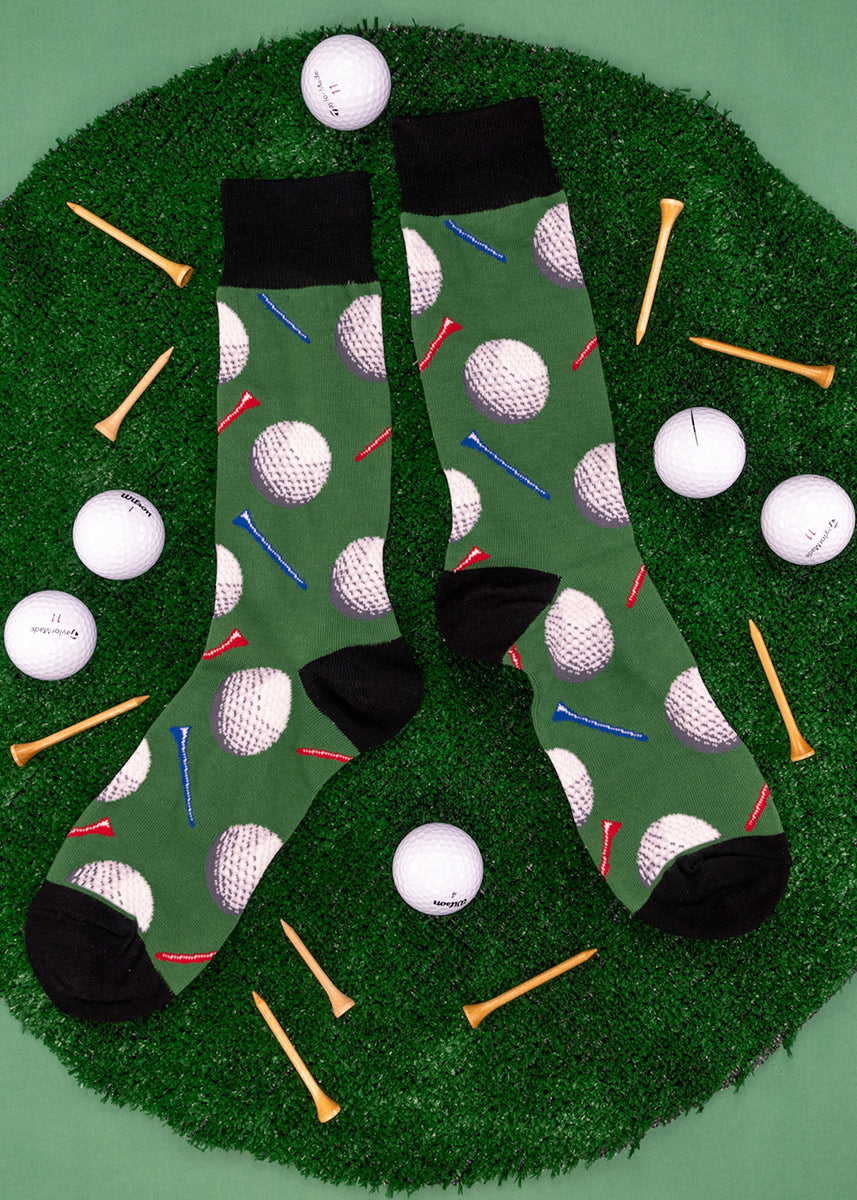 A pair of green golf-themed novelty socks lay on a circular cut of turf surrounded by golf balls and tees.