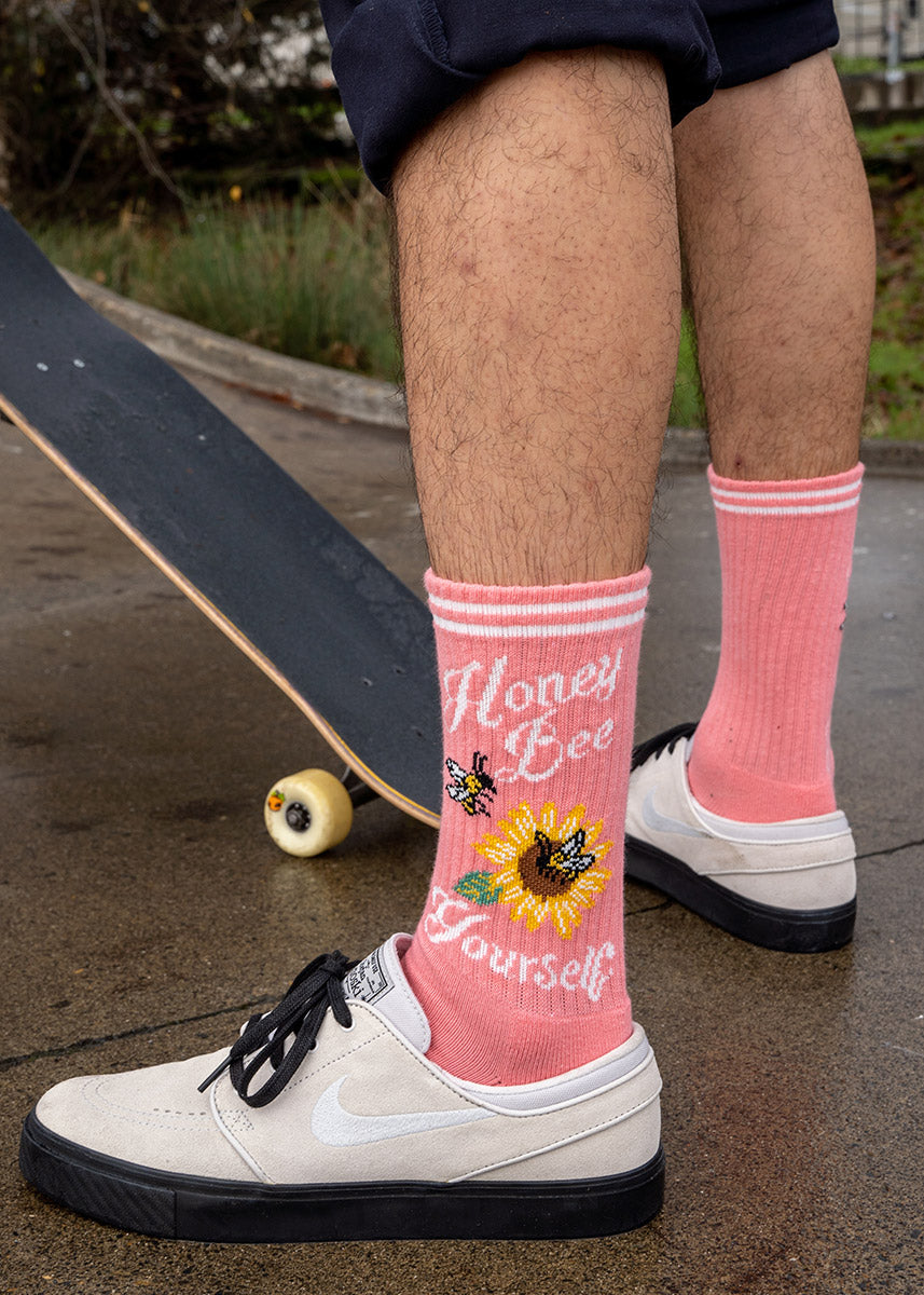 A male model wearing bee-themed novelty socks and white sneakers poses with a skateboard.