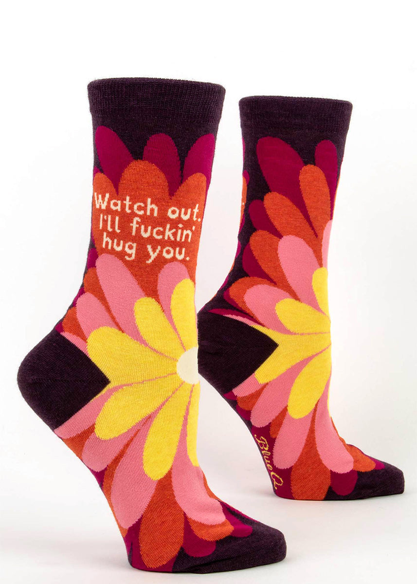 Funny swear word socks for women say, "Watch out, I'll fuckin' hug you," with a giant flower design in yellow, pinks, and orange.