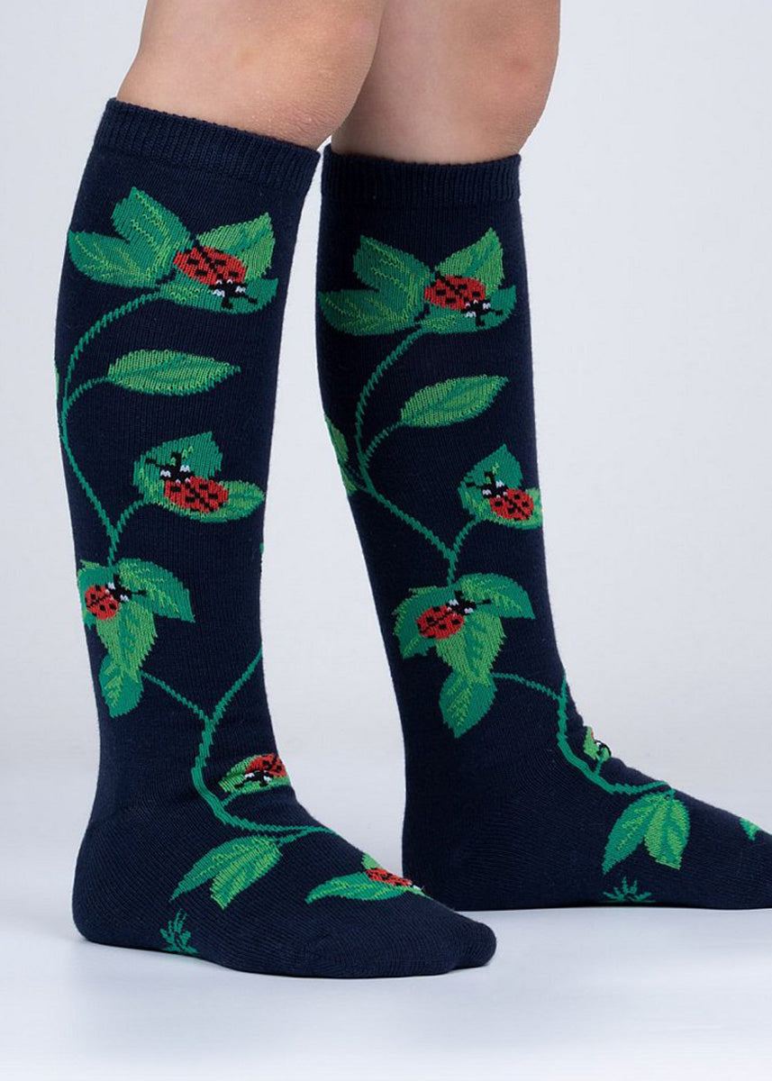 Knee-high ladybug socks for kids in dark teal show cute red and black spotted beetles roaming up and down a leafy vine.
