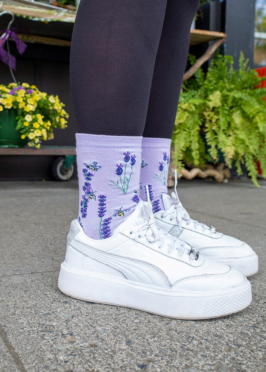 A female model wearing lavender and bee novelty socks poses on the sidewalk with plants in the background.