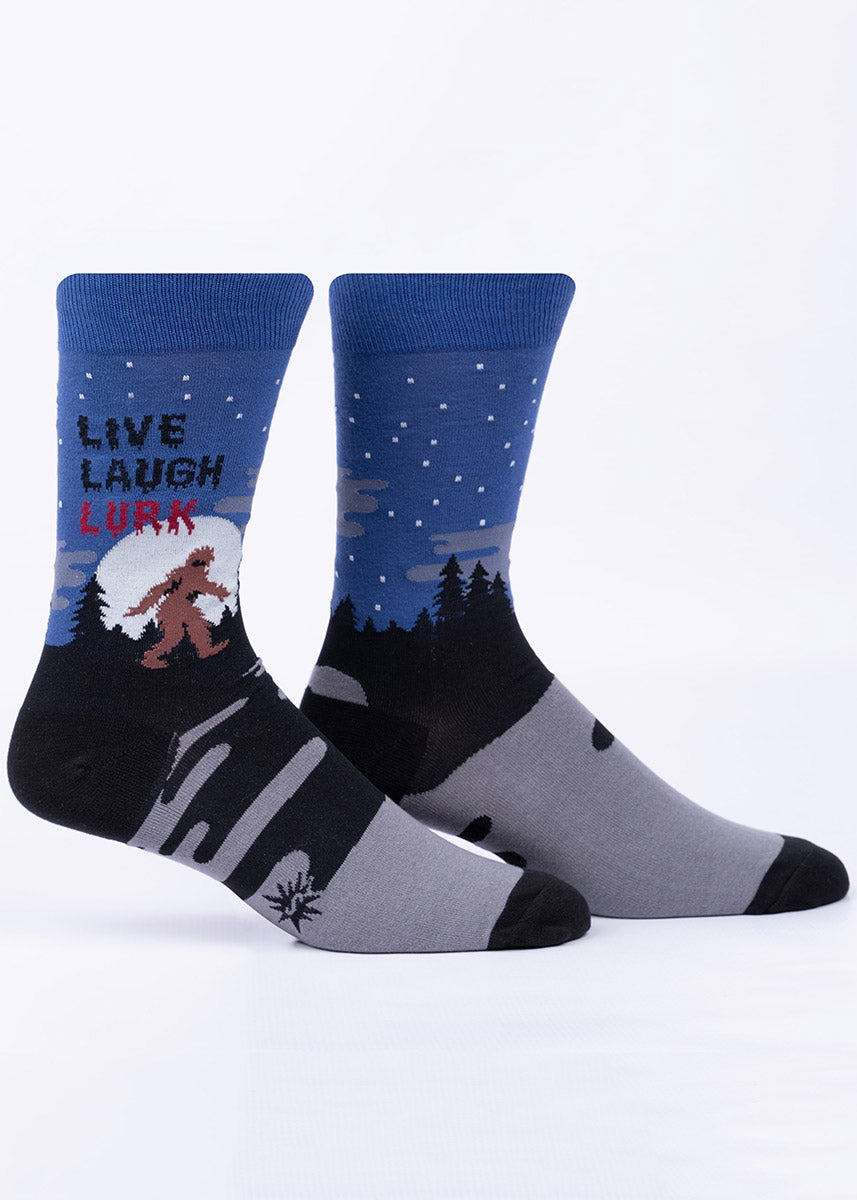 Men&#39;s crew socks in blue, black and gray say &quot;Live, Laugh, Lurk&quot; and feature Bigfoot lumbering through the woods in front of a giant glow-in-the-dark full moon.