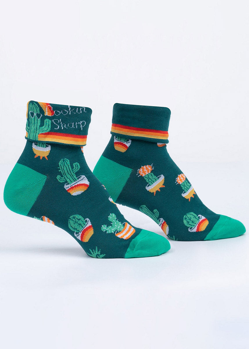 Cute green socks with rainbow stripe accents feature a cuff that can be turned down to reveal a cactus wearing sunglasses with the words, “Lookin' Sharp.”