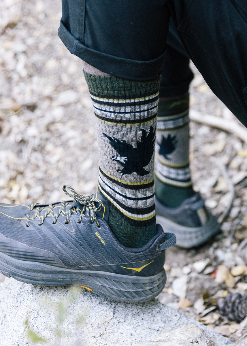 A model wearing eagle-themed wool socks and hiking shoes poses outside on a rock.