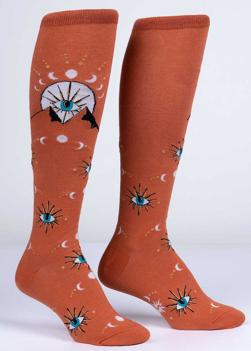 Glow-in-the-dark knee high socks feature mystic mountains, all-seeing eyes, and crescent moon symbols on a warm orange background. 