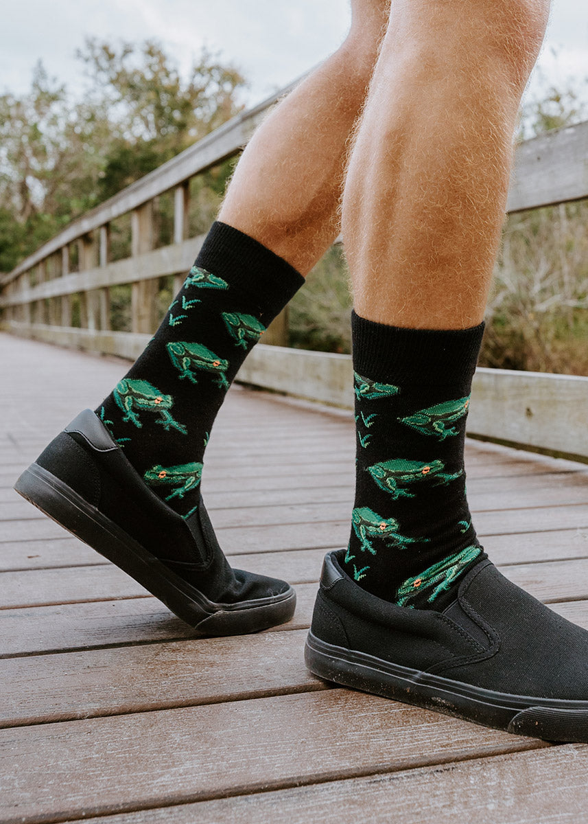 A male model wearing frog-themed novelty socks and black sneakers poses outside on a bridge